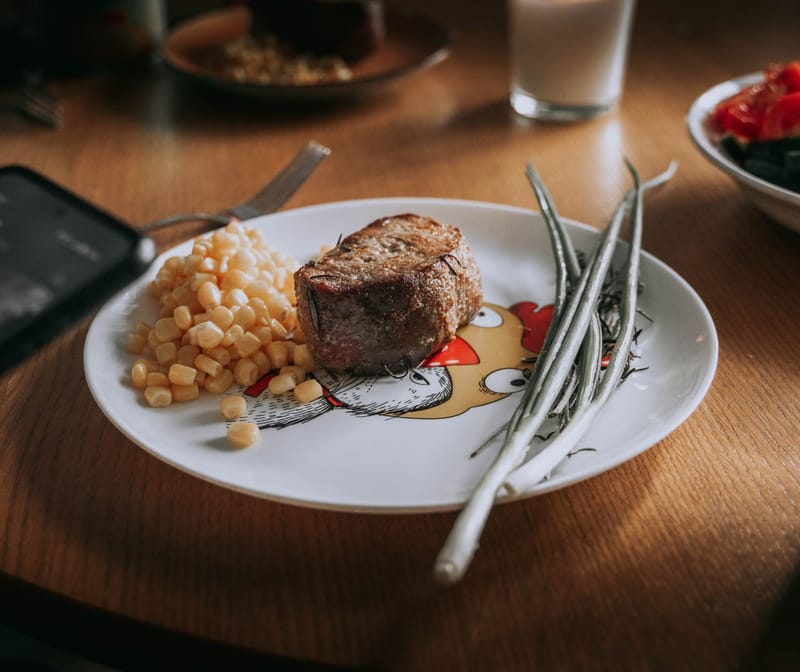 A cooked steak on a plate with green beans and corn, on a family dinner table